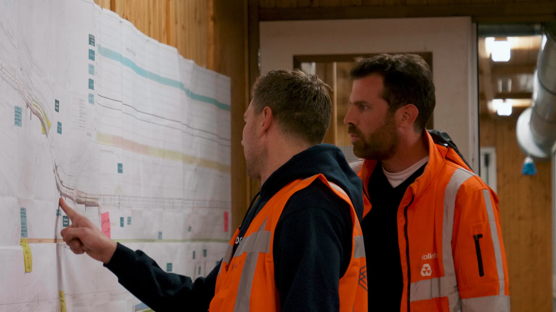 Twee ingenieurs bespreken tekeningen en planning aan de wand van een bouwkeet