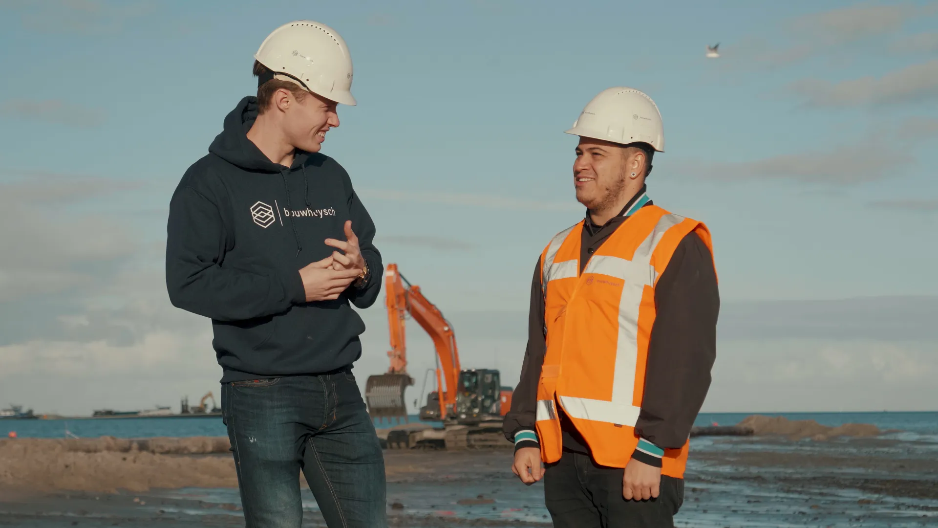 Twee jonge vakmensen in gesprek op een kustproject, met graafmachine op de achtergrond